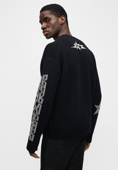 Černý pletený svetr s dlouhými rukávy. Obsahuje bílé vyšívané detaily a geometrické vzory podél rukávů a hvězdné motivy na zádech.