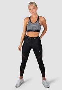 Juoda ir pilka sportinė liemenėlė su plačiu diržu ir logotipu, derinama su juodomis legginėmis, turinčiomis tinklelio išpjovas. Modelis vilki baltus sportinius batus.