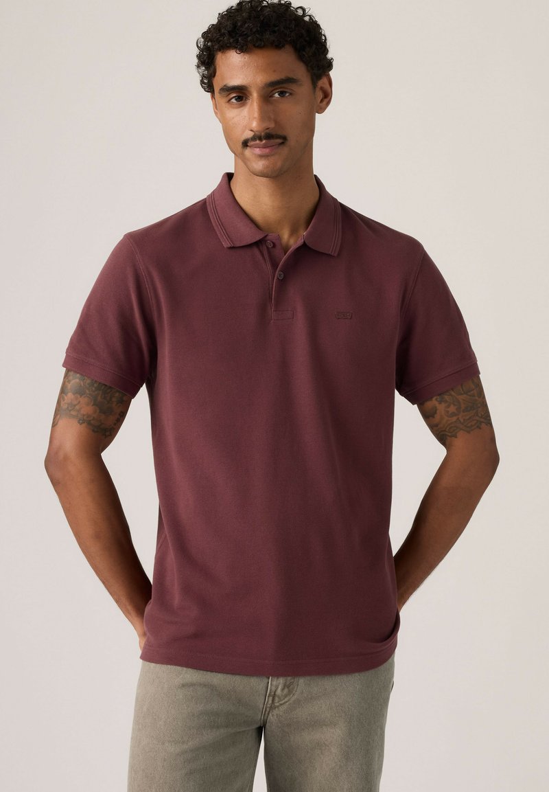 Hombre con cabello rizado y bigote lleva una camiseta polo granate y pantalones gris claro, de pie con las manos detrás de la espalda sobre un fondo liso.