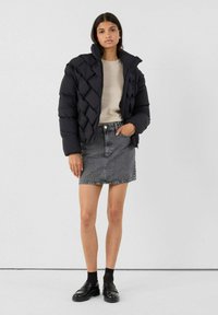 Vrouw in een zwarte gewatteerde pufferjack, beige top, grijze spijkerrok, zwarte sokken en zwarte instappers, staande tegen een effen witte achtergrond.