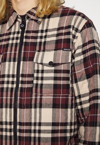 Camicia a quadri con un mix di motivi beige, burgundy e neri, con zip frontale e due tasche sul petto. Tessuto morbido.