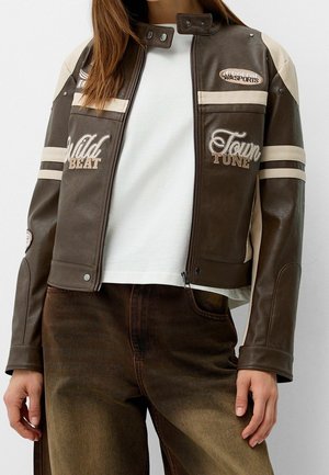 Frau trägt eine braune und beige Lederjacke mit den Aufschriften "Wild Beat" und "Town Tune", kombiniert mit einer braunen Hose und einem weißen Hemd.