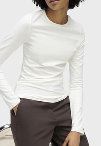 Person wearing a fitted white long-sleeve top and dark brown pants, sitting with one hand in pocket against a plain light background.