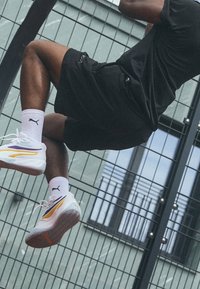 Baskets Puma blanches avec des accents jaunes et violets, en matériaux maille et caoutchouc, avec des lacets, une semelle texturée et des détails de logo.