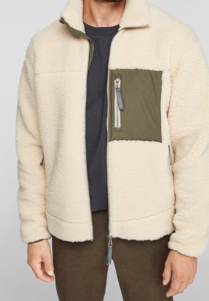 Mann trägt eine cremefarbene Fleecejacke mit olivgrünem Reißverschluss-Brusttasche, dunklem Hemd und brauner Hose, steht vor grauem Hintergrund.