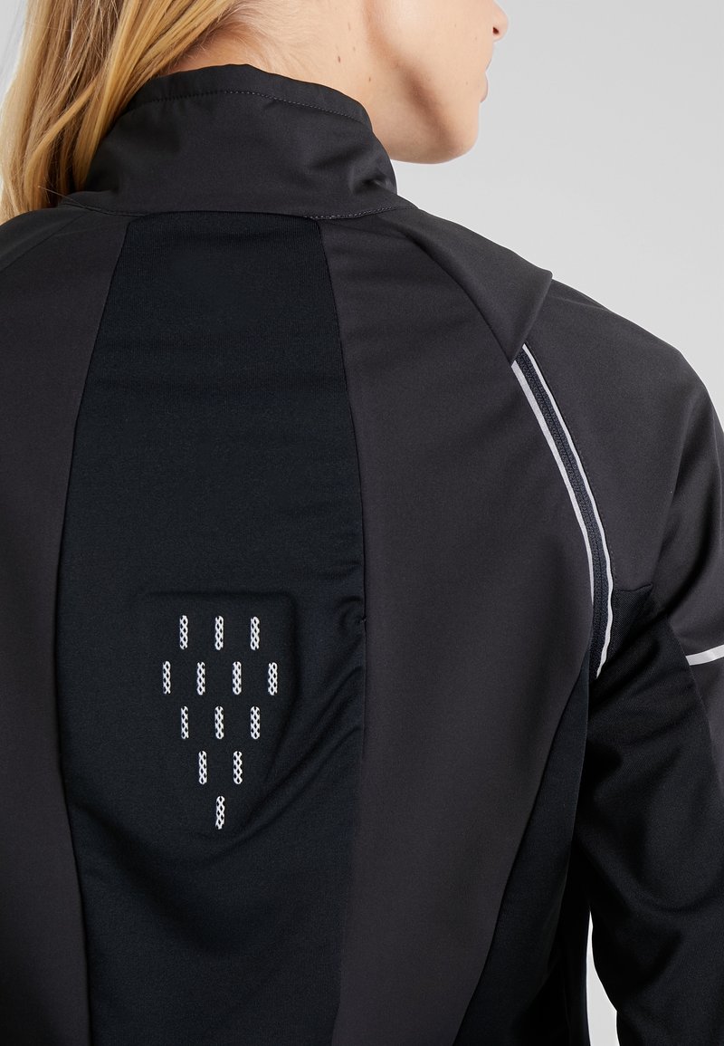 Femme portant une veste noire avec des bandes réfléchissantes sur l'épaule et un panneau en tissu ventilé sur le haut du dos.