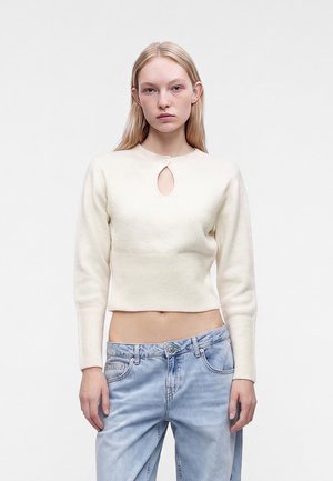 Jeune femme aux longs cheveux blonds portant un pull court crème avec un détail œillet et un jean taille haute bleu clair sur un fond uni.
