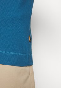 Blå långärmad skjorta med struktur och en liten orange "BOSS"-etikett vid fållen, kombinerad med ljusbeige byxor.