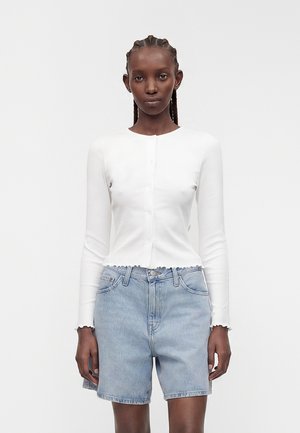 Kvinde med flettet hår iført en hvid langærmet knapbluse og lyseblå denimshorts mod en ensfarvet lys baggrund.