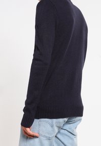 Pull en maille bleu marine avec tissu texturé, ourlet et poignets côtelés, et coupe ajustée. Présenté de dos, coupe légèrement décontractée.