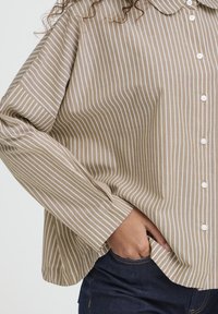 Person med krøllet hår iført en beige og hvid vertikalt stribet skjorte, med den ene hånd stukket ind i lommen på mørkeblå jeans.