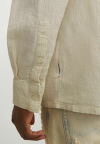 Bež tekstuuriga särgi varrukas, millel on kaks nööpi ja silt tekstiga "ORIGINALS" kõrval; kantud narmendavate denim-šortidega.