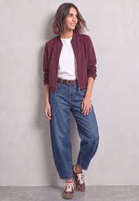 Femme portant une chemise blanche, une veste bordeaux, un jean bleu et des baskets bordeaux, debout et souriante devant un mur clair et uni.