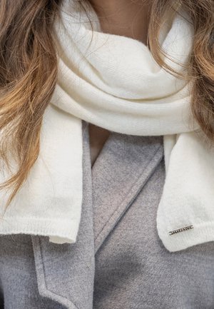 Femme portant une écharpe en laine blanche enveloppée autour du cou, par-dessus un manteau gris, avec des cheveux bruns lâches tombant sur les épaules.
