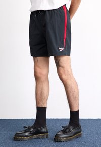 Černé šortky s červeným bočním pruhem a logem Reebok, vyrobené z lehkého materiálu. Nosič má na sobě černé ponožky a boty s silnou podrážkou.