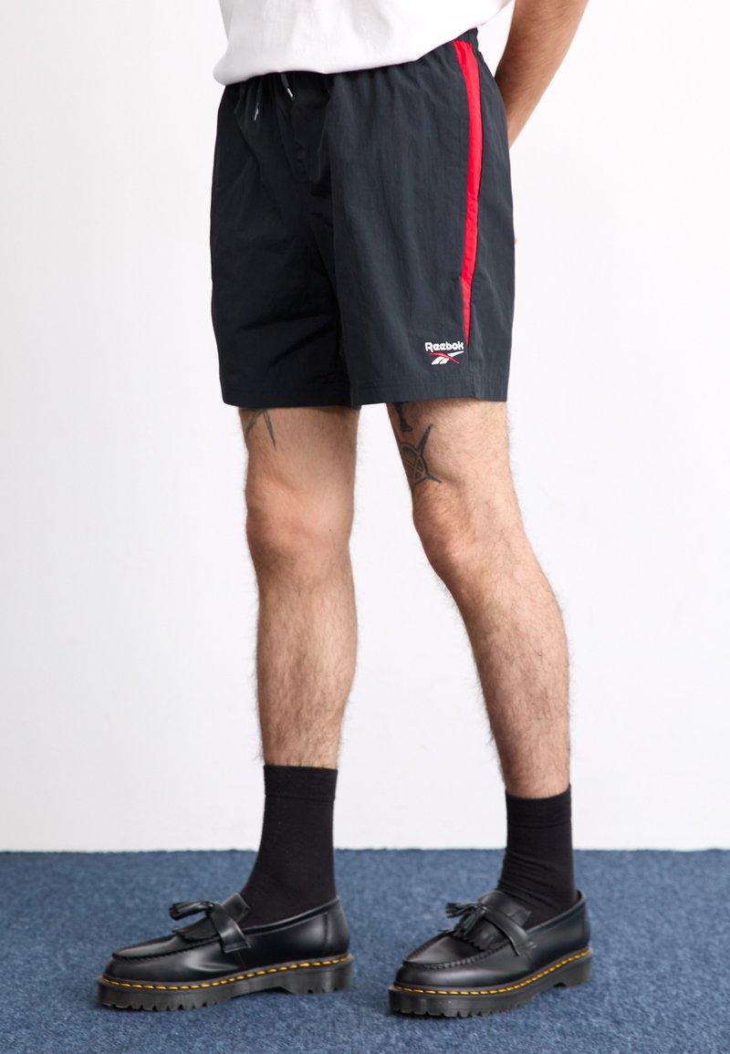Černé šortky s červeným bočním pruhem a logem Reebok, vyrobené z lehkého materiálu. Nosič má na sobě černé ponožky a boty s silnou podrážkou.