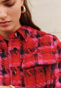 Giacca con motivo rosso e rosa, con una trama testurizzata, dotata di dettagli neri e bottoni a pressione neri lucidi; include un dettaglio di tasca frontale.