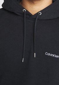 Felpa nera in tessuto misto cotone, con cappuccio regolabile con coulisse e logo "Calvin Klein" ricamato in bianco sul petto.