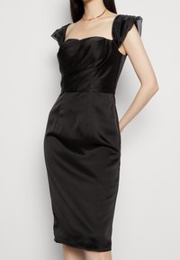 Černé saténové šaty s strukturovaným korzetem, drapovanými detaily a průhlednými, nadýchanými rukávy. Úzký střih s délkou ke kolenům.