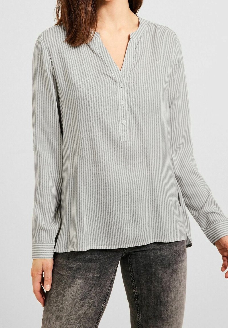 Femme portant un chemisier à manches longues gris clair à rayures verticales avec une patte de boutonnage partielle et un jean noir délavé.