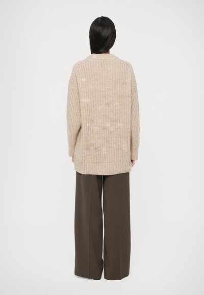 Beige gestrickte Strickjacke mit lockerer Passform, die ein strukturiertes Gewebe und verlängerte Ärmel aufweist, kombiniert mit weit geschnittenen, dunkelgrünen Hosen.