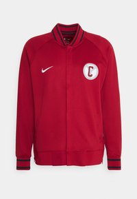 Rød jakke med høj krave, lynlås foran og en enkelt lomme. Har et rundt logo med bogstavet "C" og kontrastfarvede stribede manchetter.