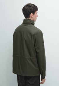 Giacca verde scuro leggera con collo alto, caratterizzata da una texture liscia e un design senza cuciture; orlo posteriore più lungo e polsini regolabili.