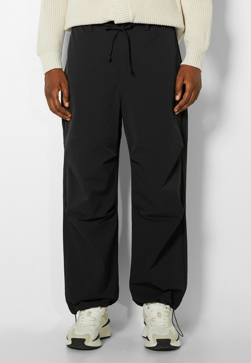 Personne portant un pantalon noir à cordon et des baskets blanches à semelles épaisses, debout devant un fond clair uni.