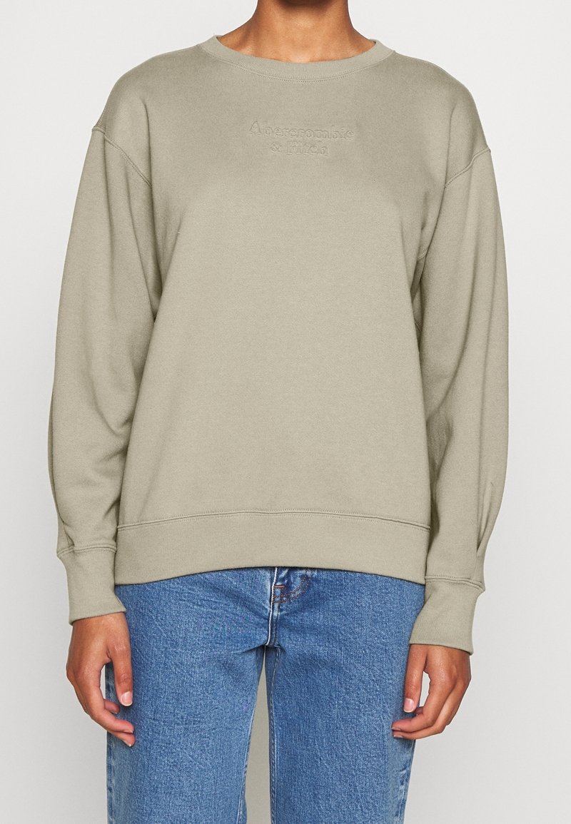 Hellgrüner Pullover mit gerippten Bündchen und Saum, mit eingeprägtem Logo auf der Vorderseite, lockerer Schnitt, aus weichem Baumwollmaterial. Kombiniert mit blauen Jeans.
