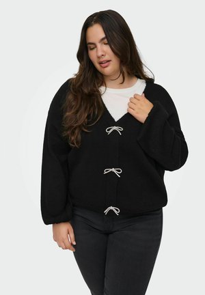 Mujer con cabello largo y castaño que lleva un cárdigan negro con tres lazos atados sobre una camisa blanca y pantalones oscuros, posando frente a un fondo sencillo.