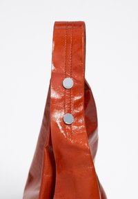 Bolso de cuero naranja y brillante con una correa ancha y acentos de botón plateado. Presenta una superficie texturizada y una forma elegante y moderna.