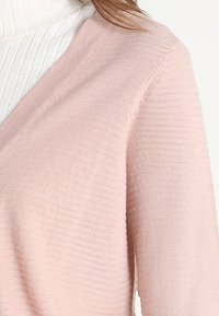 Ljusrosa cardigan med V-ringning, ribbat mönster och mjukt stickat tyg, över en vit ribbad polotröja.