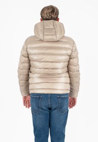 Persoon met kort haar draagt een beige gewatteerde jas met capuchon en blauwe jeans, met de rug naar de camera gekeerd tegen een witte achtergrond.