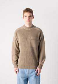 Young man wearing a beige knitted sweater with chest pocket and light blue jeans, standing against a plain white background.