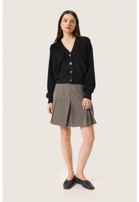 Svart stickad cardigan med v-ringning och sex knappar, tillsammans med en figursydd grå plisserad kjol. Modellen har på sig svarta texturerade skor utan häl.