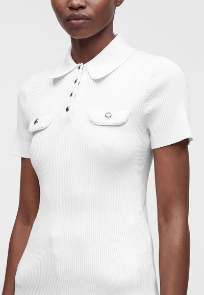 Femme portant un polo blanc à manches courtes en tissu côtelé, avec col, patte de boutonnage et deux petites poches poitrine à boutons.