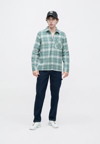 Πουκάμισο φανέλας με πράσινο καρό, μακριά μανίκια και μία τσέπη στο στήθος, σε συνδυασμό με σκούρο μπλε παντελόνι και λευκά αθλητικά παπούτσια. Μαύρο καπέλο με ρίγες.