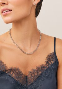 Silverfärgat pärlhalssmycke med en slät yta, bärs över en mörk spetslinne med blommig spetskant och justerbara tunna axelband.