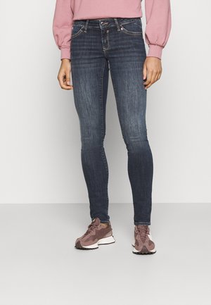Cienkie dżinsy w ciemnoniebieskim denimu z wyblakłym wykończeniem, z klasycznym pięciopocketowym designem i tonującym ściegem. Zestawione z brązowymi sneakersami.