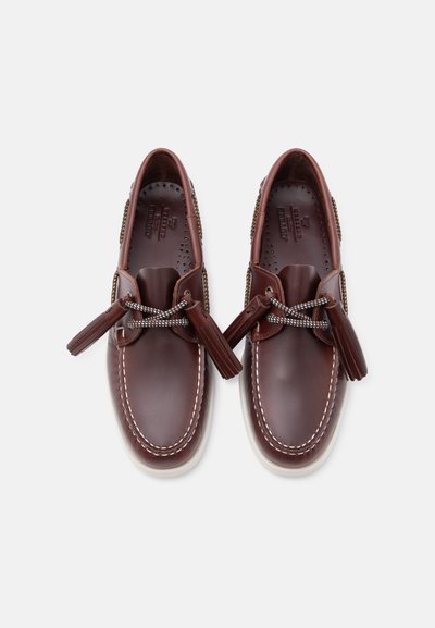 Chaussures bateau en cuir marron avec coutures blanches, lacets à motifs noués avec des pompons, et semelles perforées, vues de dessus sur fond blanc.