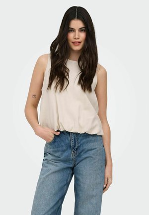 Femme aux longs cheveux foncés portant un haut beige sans manches et un jean bleu, debout avec une main dans la poche devant un fond uni.