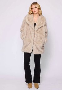 Lichtbeige faux fur jas met een brede kraag, zachte textuur, verlaagde schouders en zakken aan de voorkant, gecombineerd met zwarte nauwsluitende broek en beige hakken.