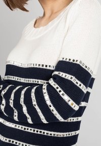 Sweter z dzianiny z naprzemiennymi białymi i granatowymi paskami oraz metalicznymi prostokątnymi akcentami, okrągłym dekoltem i prążkowanymi mankietami.