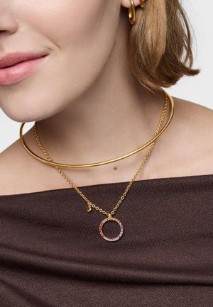 Primo piano di una persona che indossa due collane d'oro, una con un choker liscio e l'altra con un ciondolo circolare colorato su una catena.