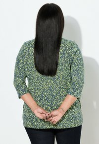 Grøn og marineblå mønstret top med 3/4-ærmer, lavet af et blødt stof, kombineret med mørke jeans. Håret er langt og glat.