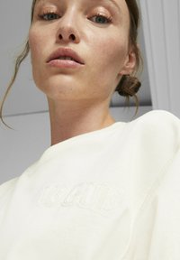 Biela mikina vyrobená z jemnej látky; má veľký, vystúpený nápis "VOGUE" na prednej strane; okrúhly výstrih a mierne voľný strih.