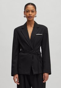 Black blazer made of smooth fabric, featuring a deep V-neck, a belted waist, long sleeves, and a front pocket with a white accent.