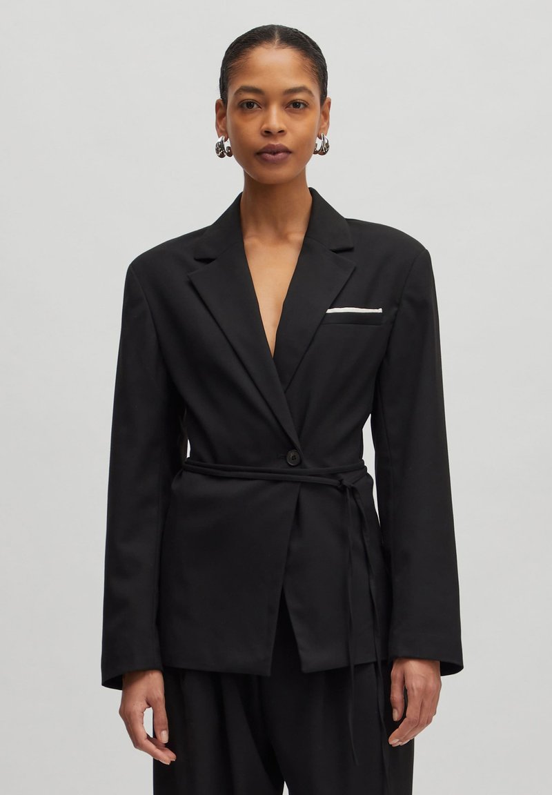 Black blazer made of smooth fabric, featuring a deep V-neck, a belted waist, long sleeves, and a front pocket with a white accent.