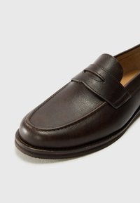 Mocasin de cuero marrón con una textura suave, punta redondeada y una correa decorativa en el empeine. Detalles de costura a lo largo de la suela.