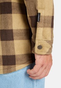 Braun- und beige-karierte Flanellshirt mit Fischgratstruktur, Druckknopf am Ärmel und einem kleinen schwarzen Etikett am Ärmel.